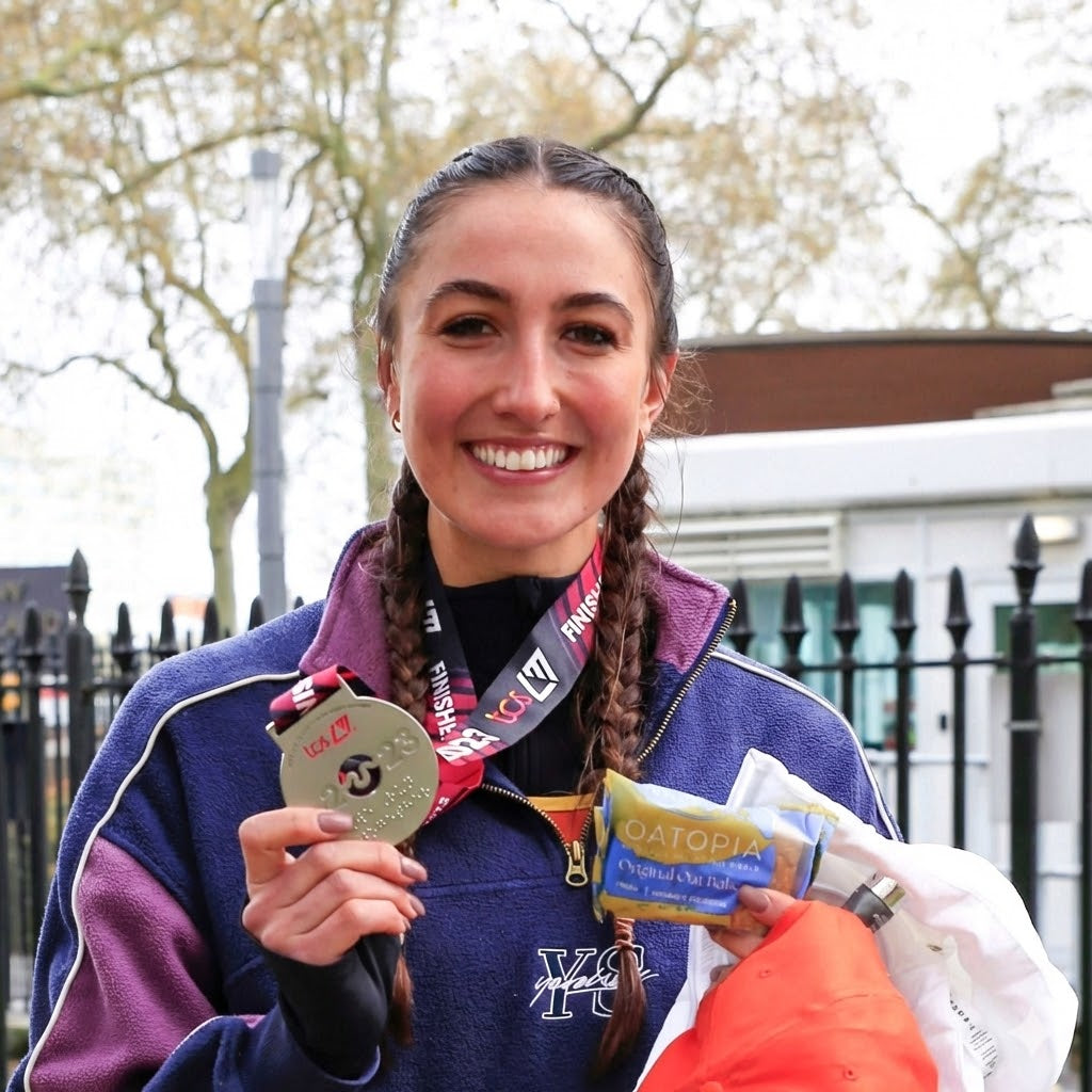  A woman proudly holds a medal in one hand and a bag of Oatopia flapjack in the other, smiling with joy.