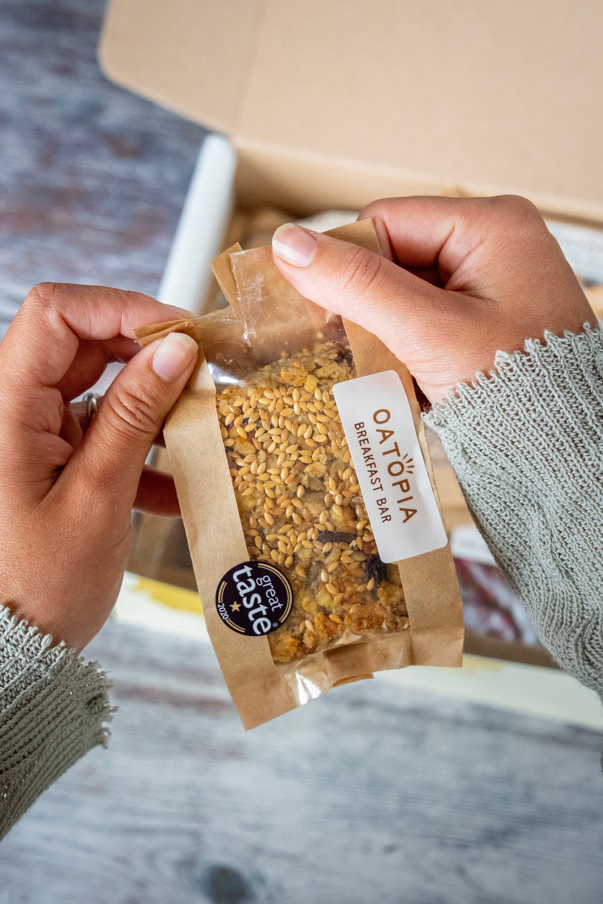 A person's hands holding a clear bag of Oatopia flapjack Bar inside a cardboard box, with a gray knit sweater sleeve visible.
