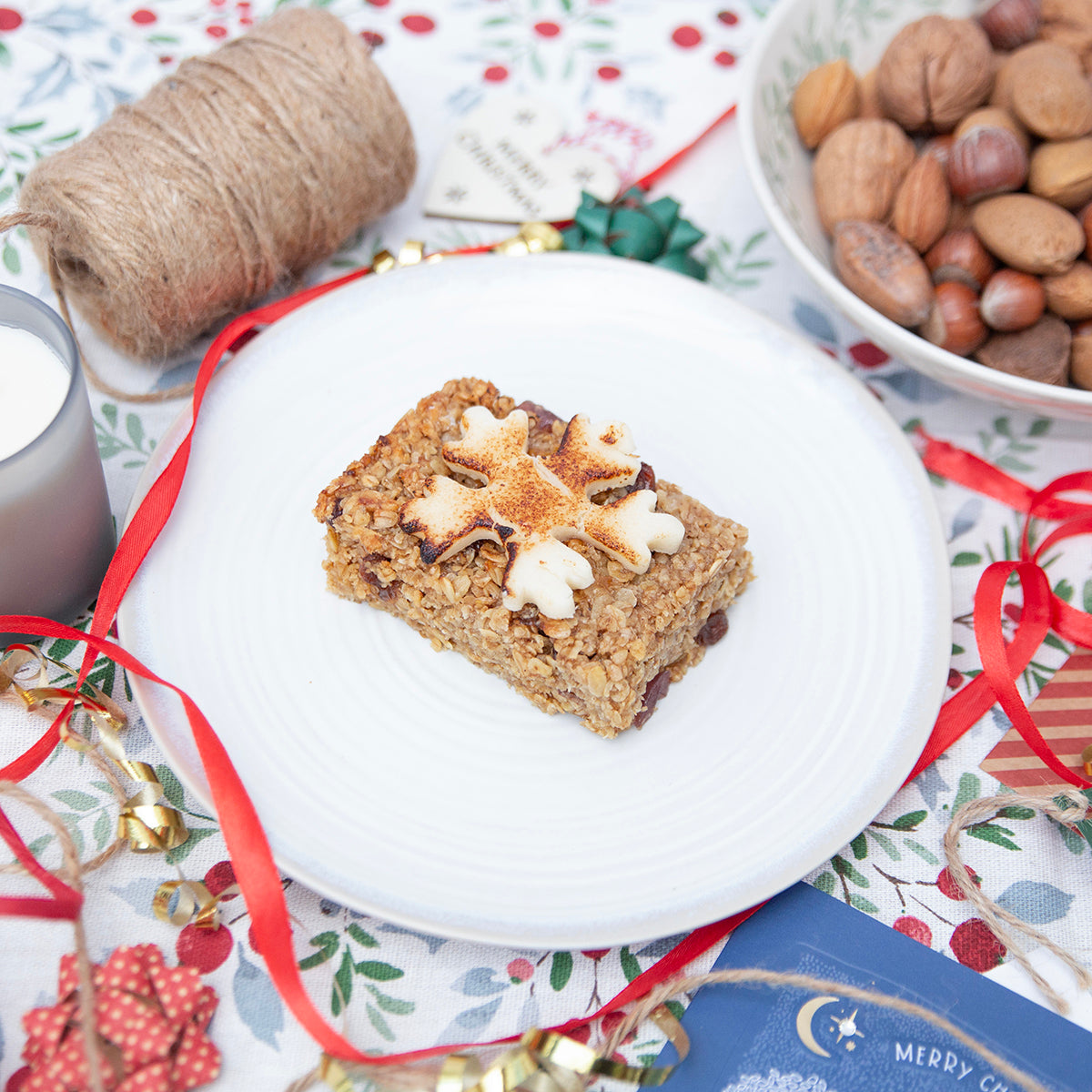 A flapjack with a marzipan topping on a white plate, surrounded by Christmas decorations including a candle, nuts, and festive cards.