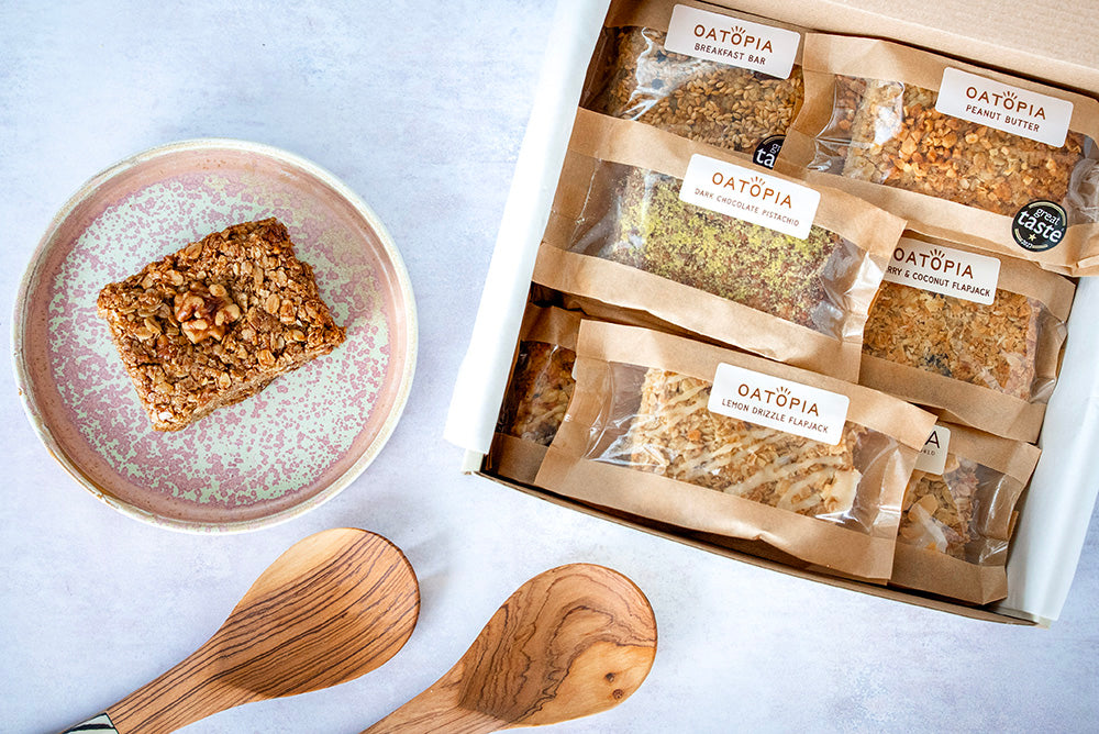 Box of wrapped Oatopia flapjack bars with a flapjack on a plate and wooden spoons alongside on a light background