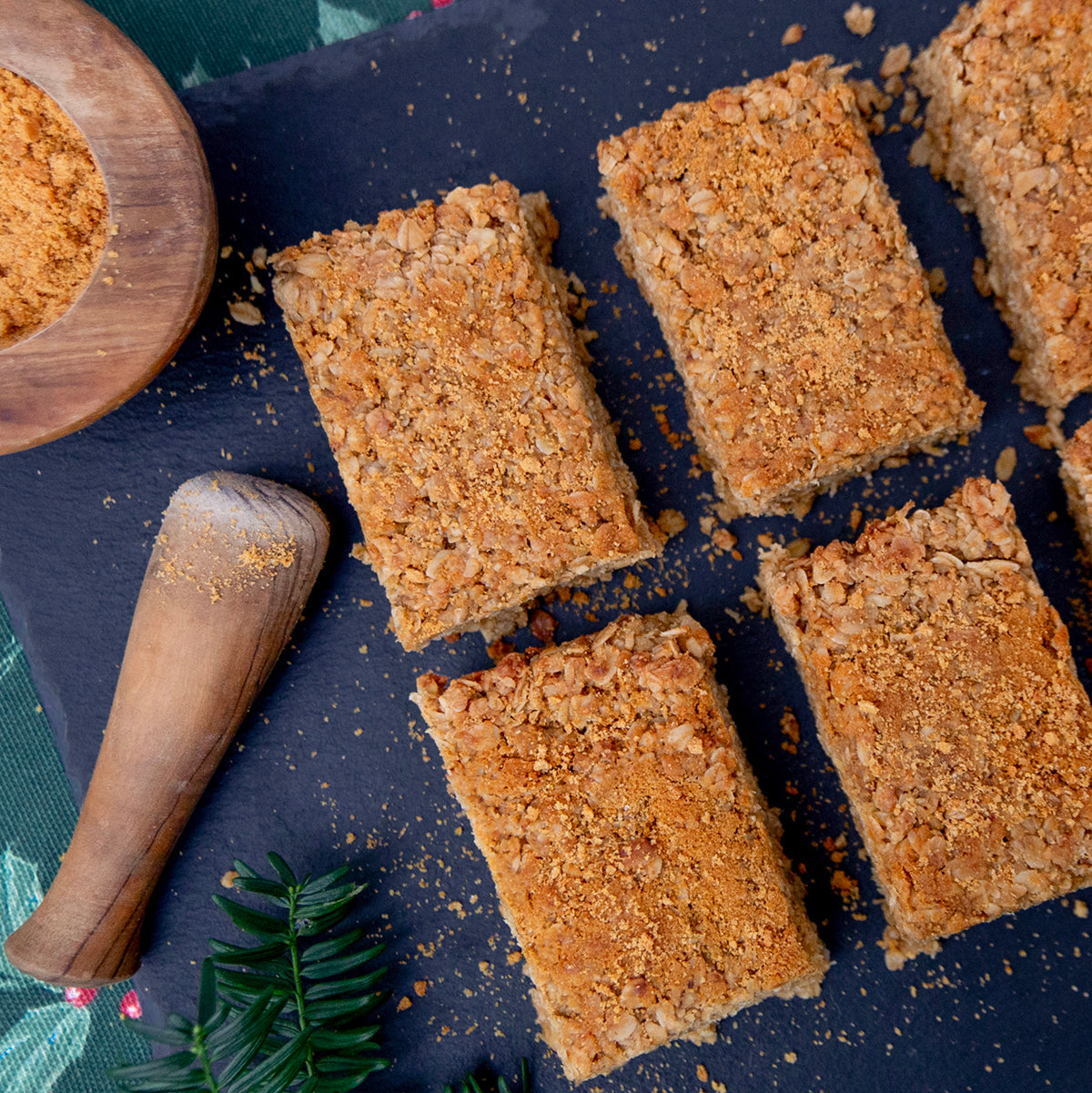 A set of gingerbread flapjacks on a dark surface, dusted with brown sugar and biscuit crumbs, with a wooden spoon in the foreground.