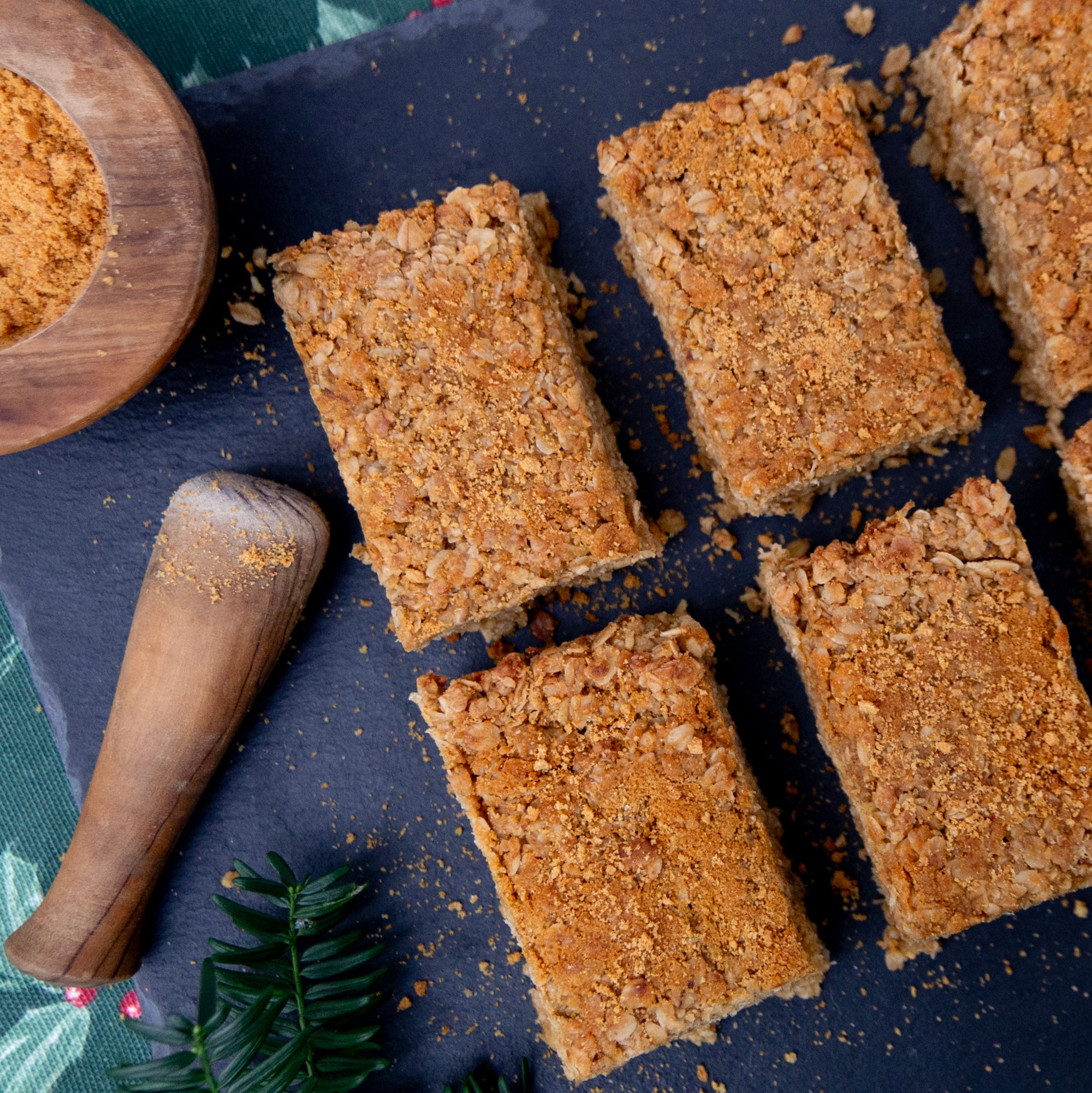 Square brown baked gingerbread flapjacks on a dark surface with a wooden bowl of spices and a pestle.