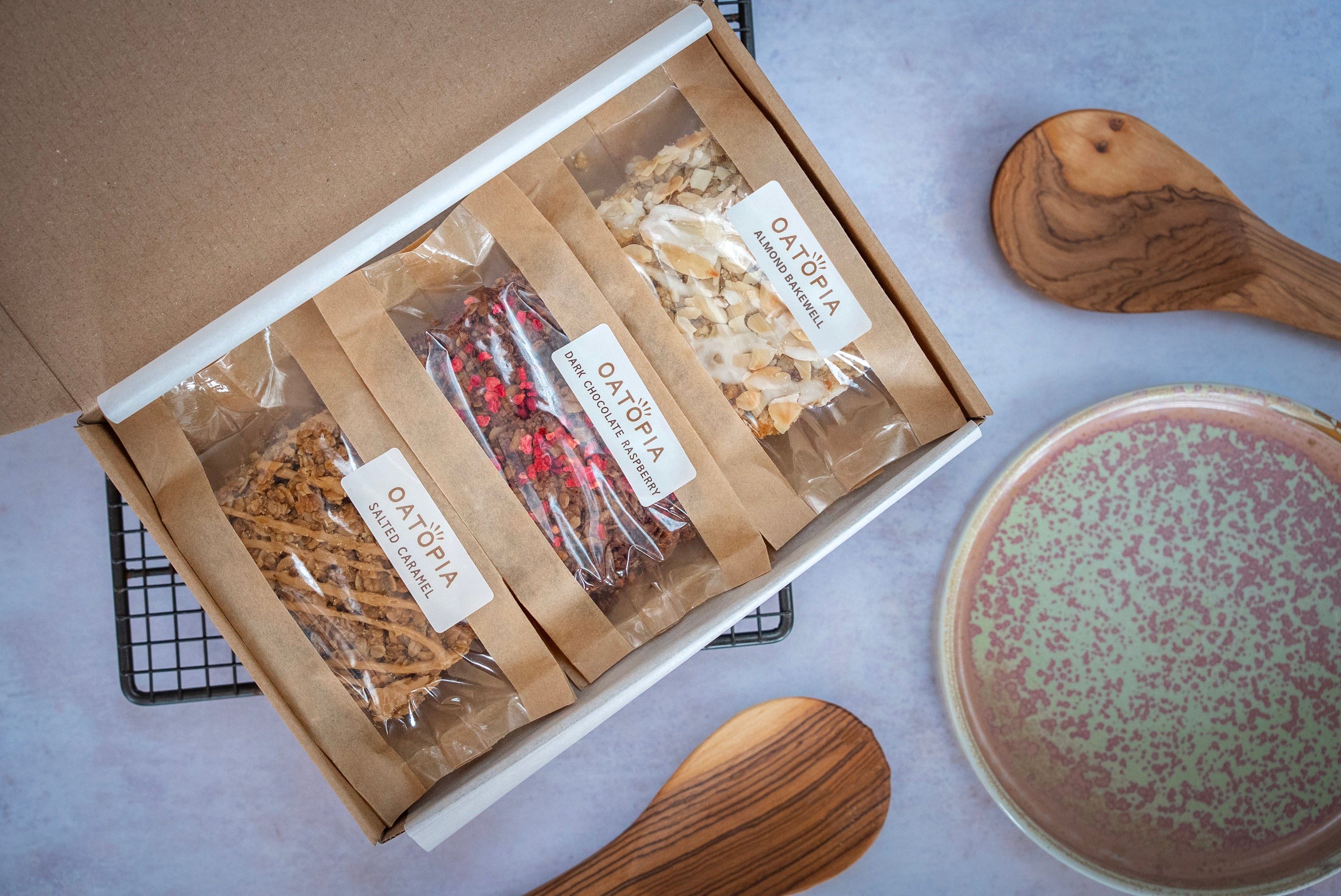 Open box containing Oatopia vegan flapjack bars on a light gray surface with a wooden spoon and decorative plate.
