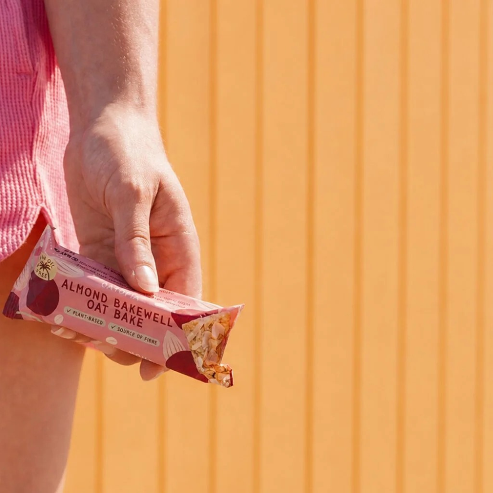 A hand holding a pink and white 'Almond Bakewell Oat Bake' snack bar against a bright orange background.