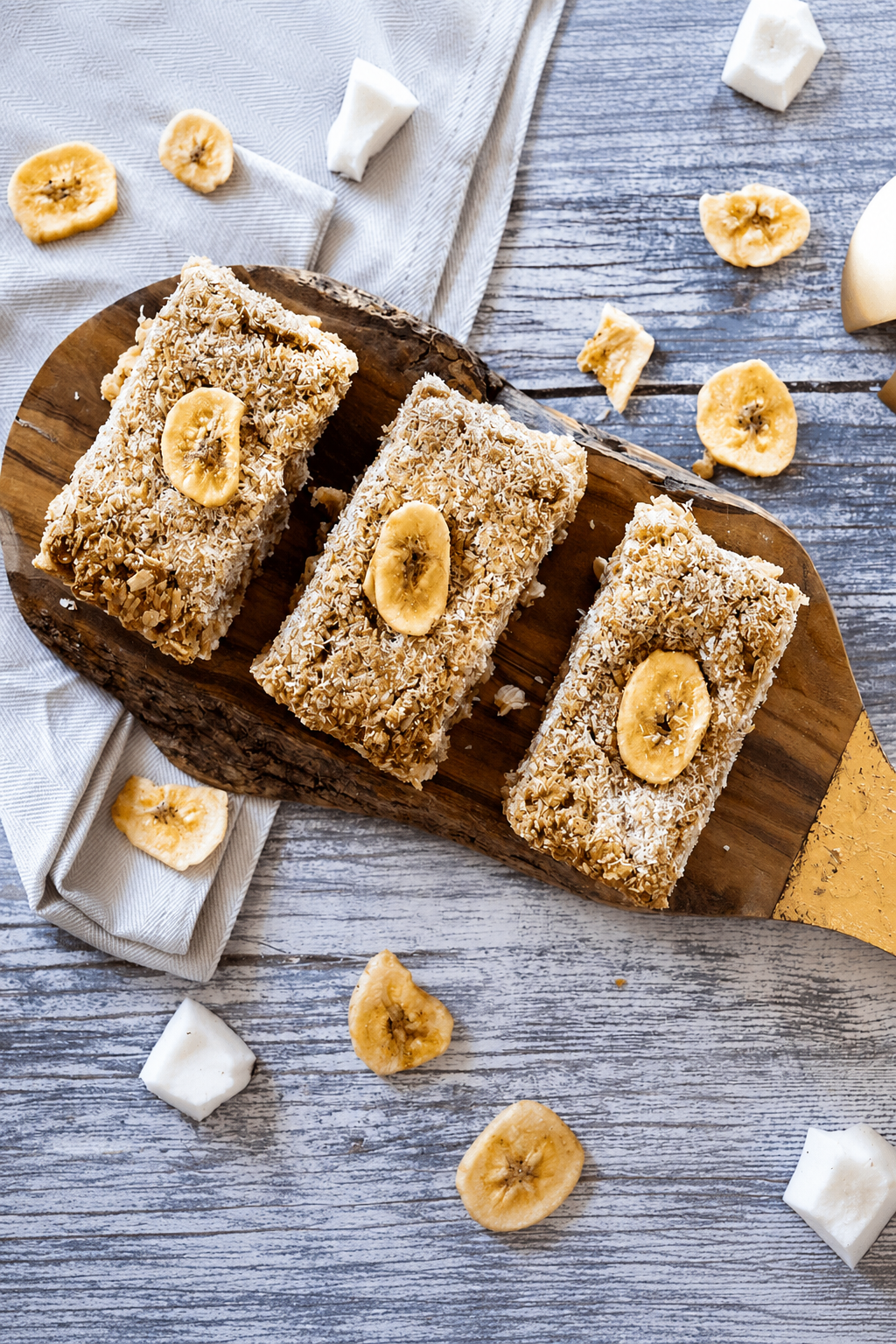 Three rectangular banana nut bars topped with banana slices, arranged on a wooden serving board over a gray rustic surface with scattered banana chips and white marshmallows.