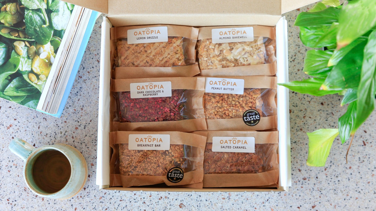 An open cardboard box containing six Oatopia granola bars in clear plastic packaging with brown paper sleeves. The box is photographed from above on a speckled countertop, surrounded by a book, a mug of coffee, and green houseplants.