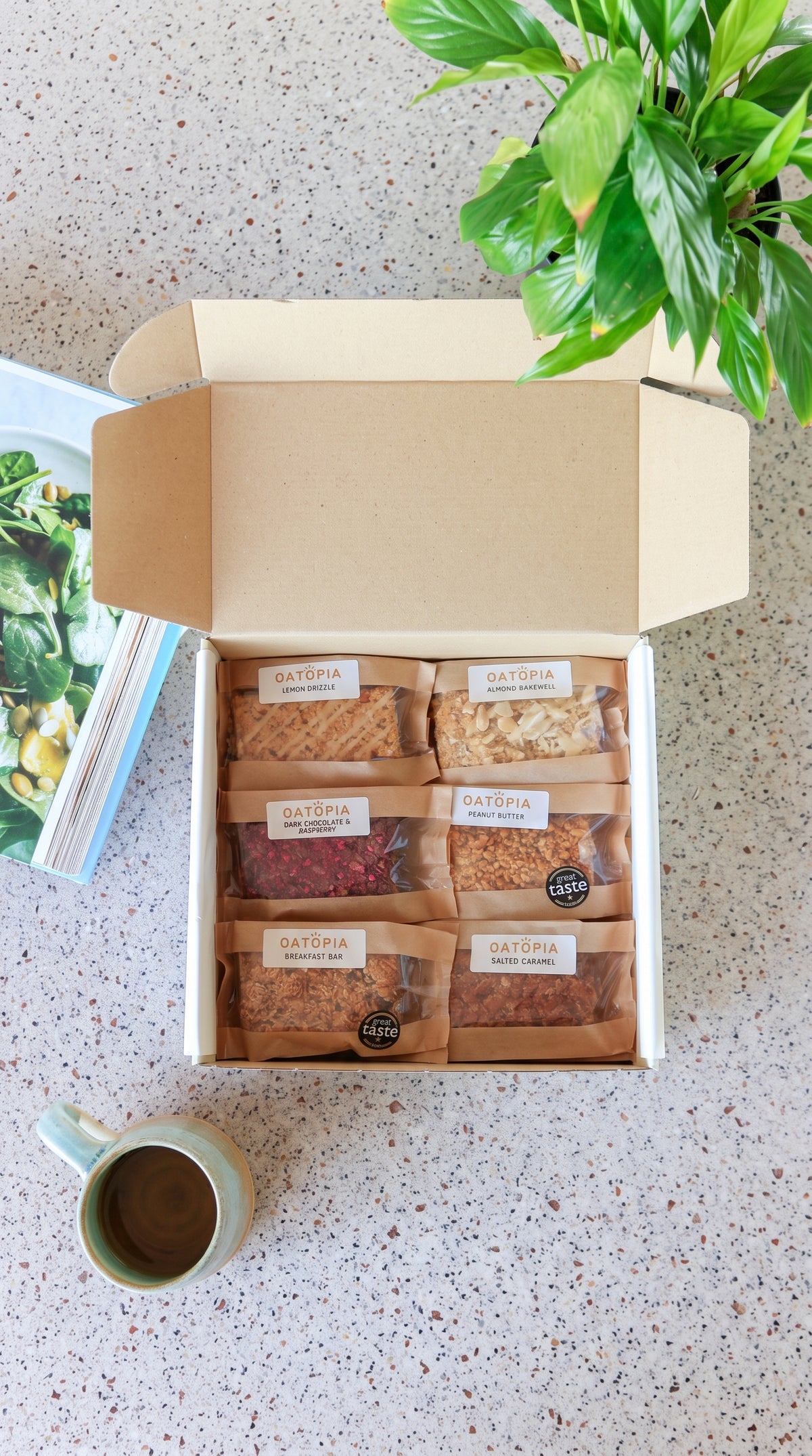 A table features a box of Oatopia_Best_Seller_collection_Gift_Box alongside a variety of snack options, inviting and ready to enjoy.