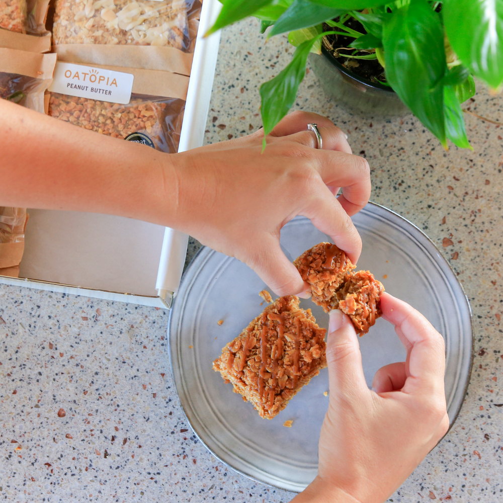 Hands breaking a soft-baked Oatopia Salted Caramel flapjack over a plate beside the gift box.