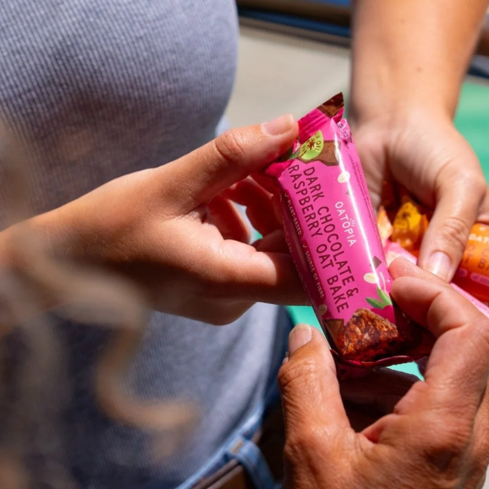  A woman smiles while holding a pink bar of Oatopia Dark Chocolate & Raspberry Oat bake.