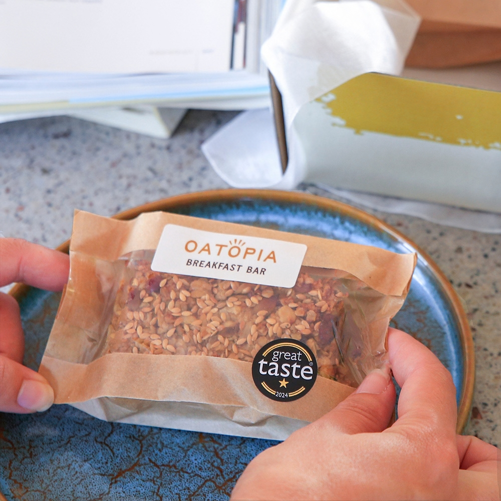 A person holds a Oatopia Breakfast Bar in a clear, brown paper sleeve on a blue ceramic plate. The bar is filled with oats, nuts, and dried fruit.