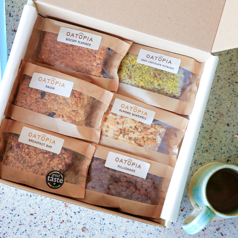 An open white box filled with six individually wrapped Oatopia breakfast items on a speckled countertop, next to a small green ceramic mug. The products include biscotti flatbread, raisin, dark chocolate pistachio, almond baklava, breakfast bar, and millomsa.