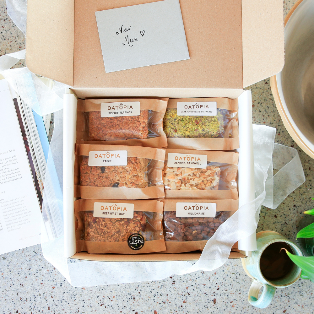 An open kraft gift box containing six individually wrapped Oatopia flapjack packs, arranged in two rows of three on white tissue paper. A handwritten note inside the lid says 'New Mom'.