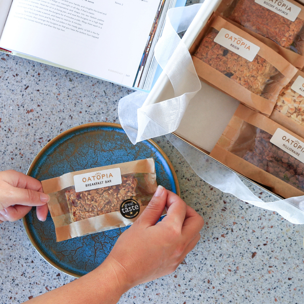 Hands holding a packaged of flapjack bar over a blue plate, with a branded box of Oatopia Breakfast Bars and an open book on a speckled countertop.