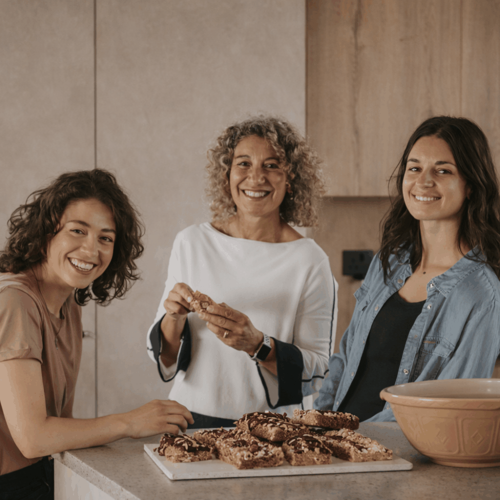In a bright kitchen, three women gather around a counter filled with food, smiling and enjoying their cooking experience.