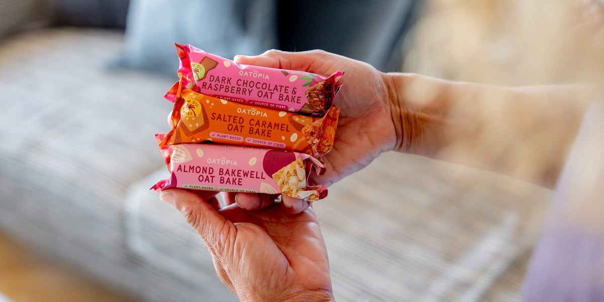 Three packaged oat bars held in two hands against a softly blurred indoor background. The wrappers are pink, orange, and coral tones with readable text including 'Dark Chocolate & Raspberry Oat Bar,' 'Salted Caramel Oat Bar,' and 'Almond Bakewell Oat Bar.
