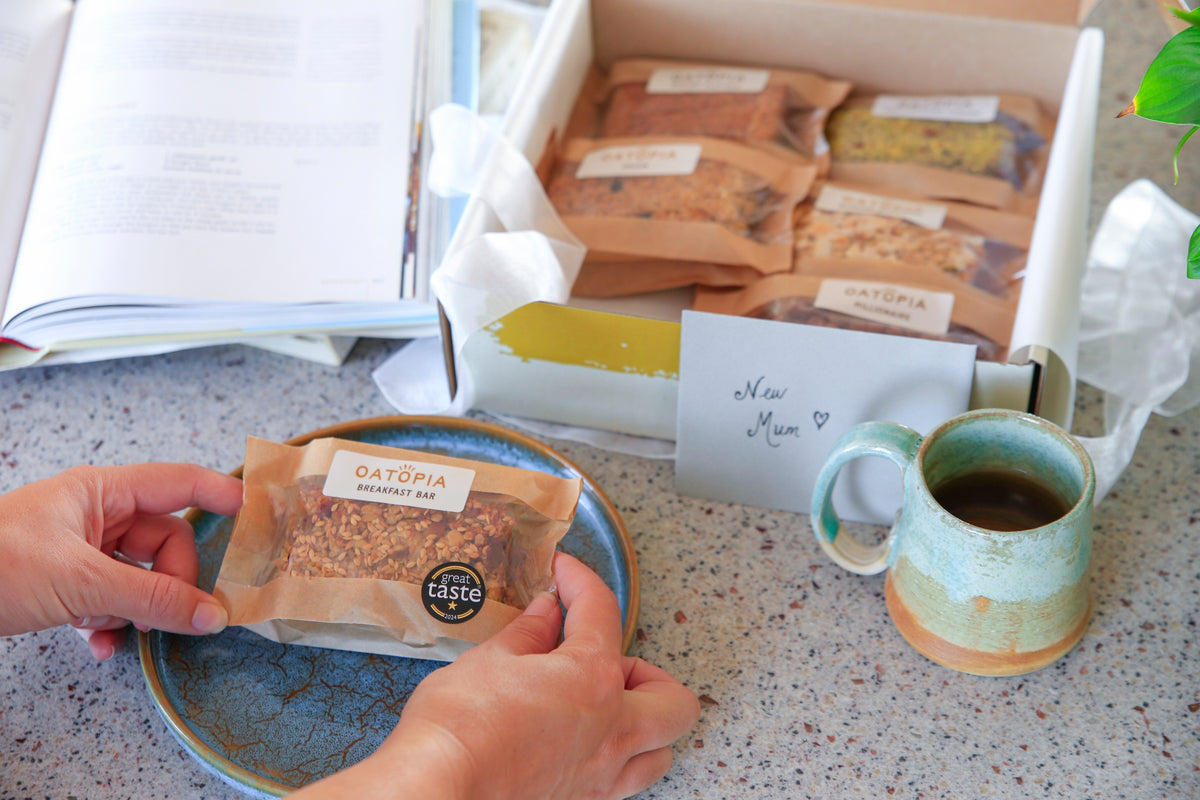 A person's hands hold a packaged Oatopia Breakfast Bar on a blue plate, with an open gift box of Oatopia bars, an open book, and a ceramic mug on a speckled countertop.