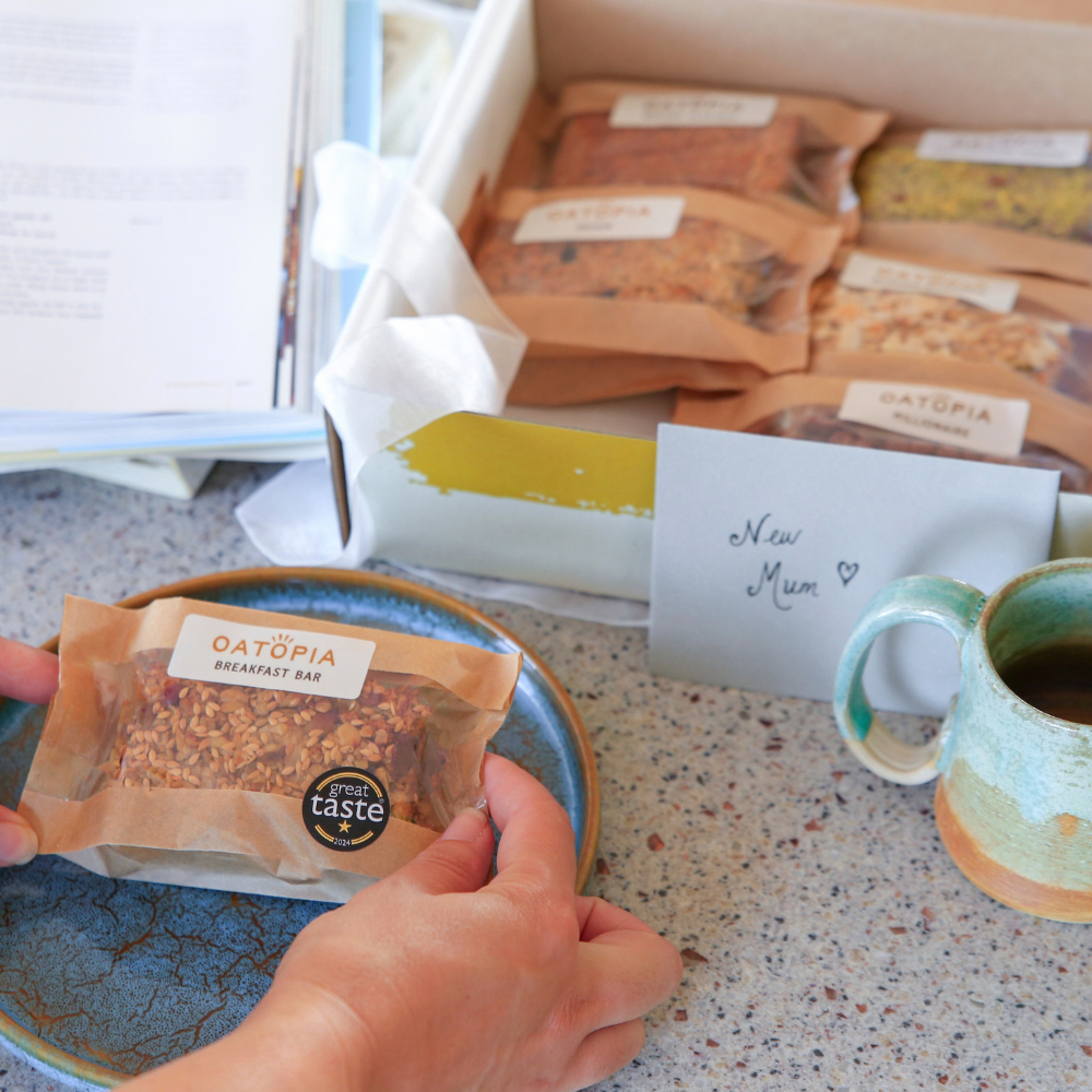A person's hands hold a oatopia breakfast bar on a blue plate, with an open box of oatmeal bars in the background and a ceramic mug on a speckled countertop.