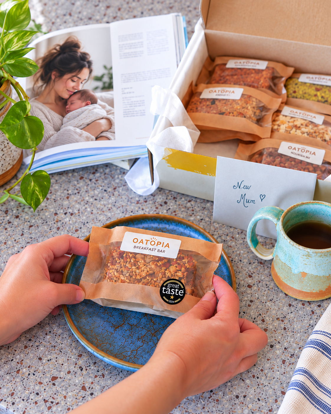 A person's hands hold a single Oatopia Breakfast Bar on a blue plate, with an open Oatopia gift box containing multiple breakfast bars in the background, alongside an open magazine, a ceramic mug, and a plant on a speckled countertop.