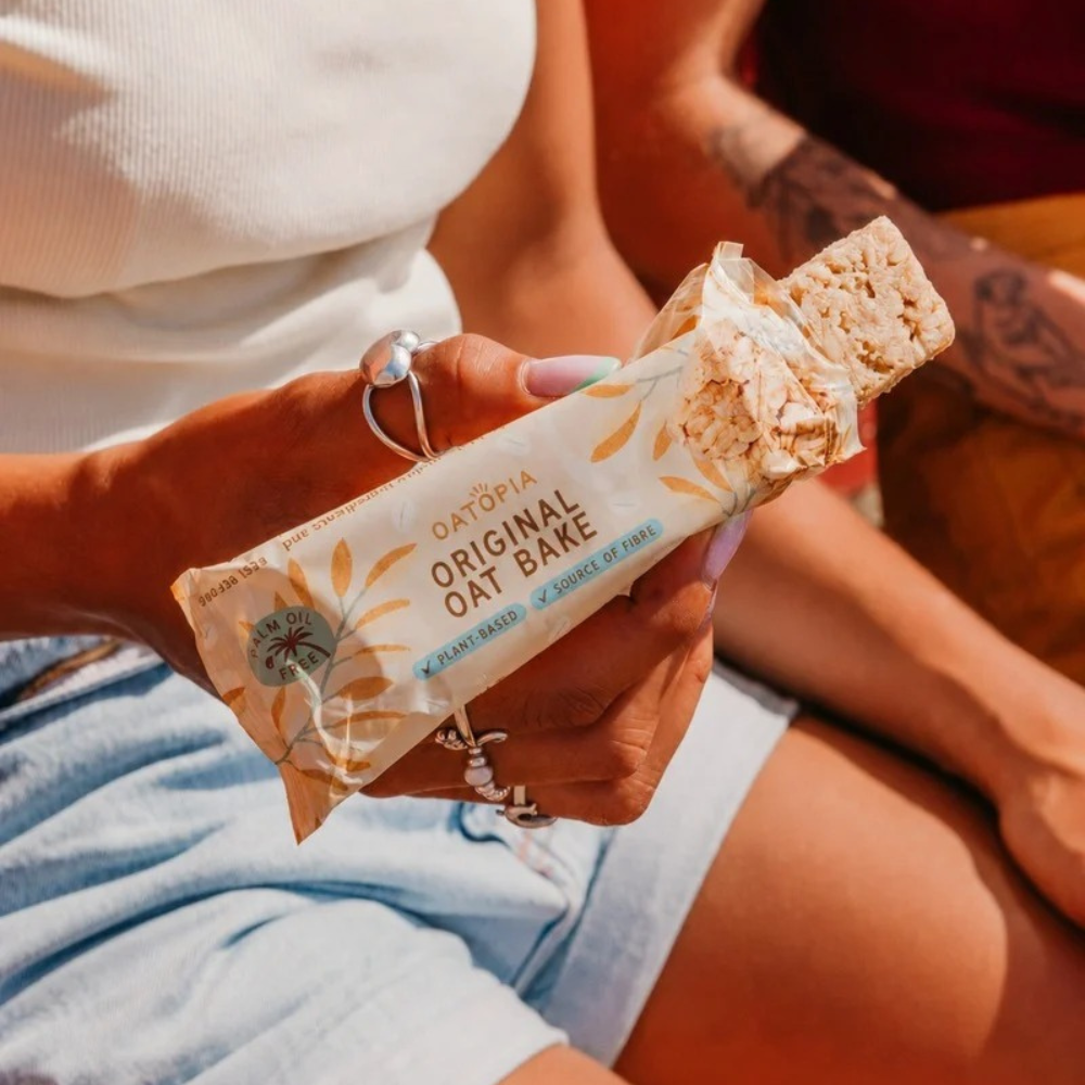 Hands holding an Oatopia Breakfast Bar over a blue plate beside the New Mum Gift Box.