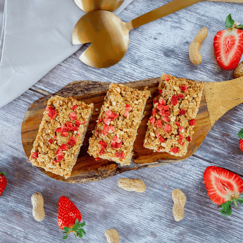 Three flapjack bars with strawberry pieces on a wooden board, surrounded by strawberries and peanuts on a gray surface.