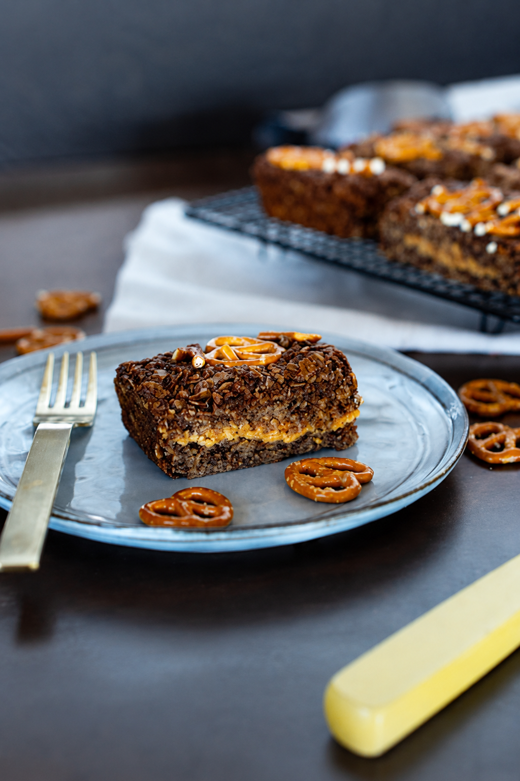 A slice of flapjack with a caramel-like filling and pretzel pieces on top, served on a blue plate with a fork, with more cake on a cooling rack in the background.
