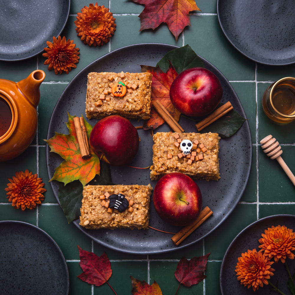 Oatopia Four Halloween-themed flapjacks arranged on a dark grey plate with red apples, cinnamon sticks, and autumn leaves, set on a green tiled table with a teapot and plates.