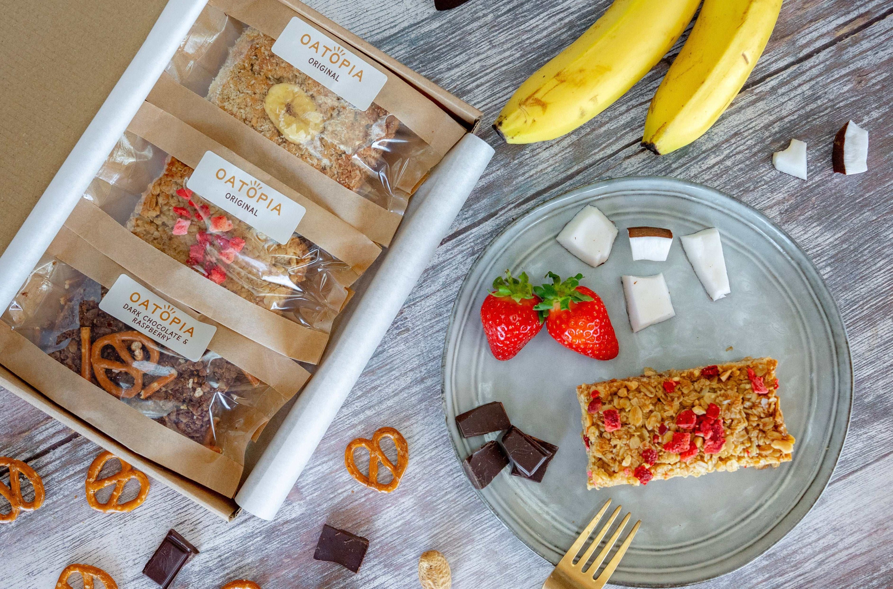 Oat bars from Oatoria on a plate with bananas, strawberries, and pretzels on a wooden surface.