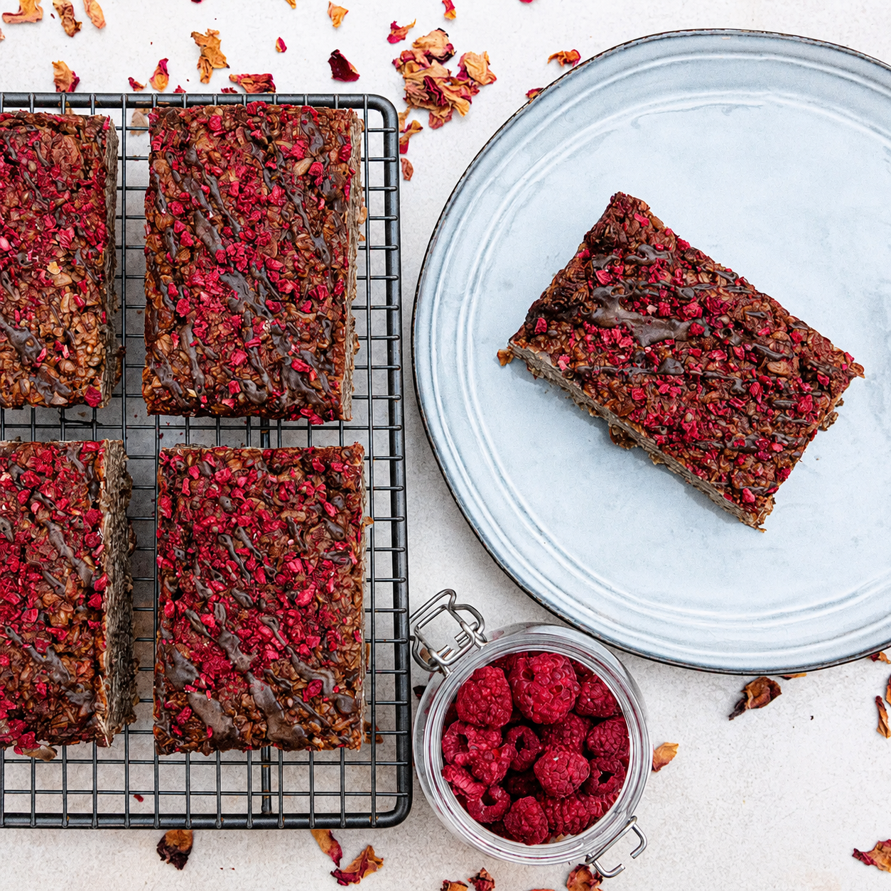 Four rectangular chocolate raspberry flapjack bars sit on a cooling rack. They are topped with dried raspberries and drizzled with chocolate. A plated slice sits beside the rack. A small jar of fresh raspberries sits on the light surface. Dried raspberries are scattered around.