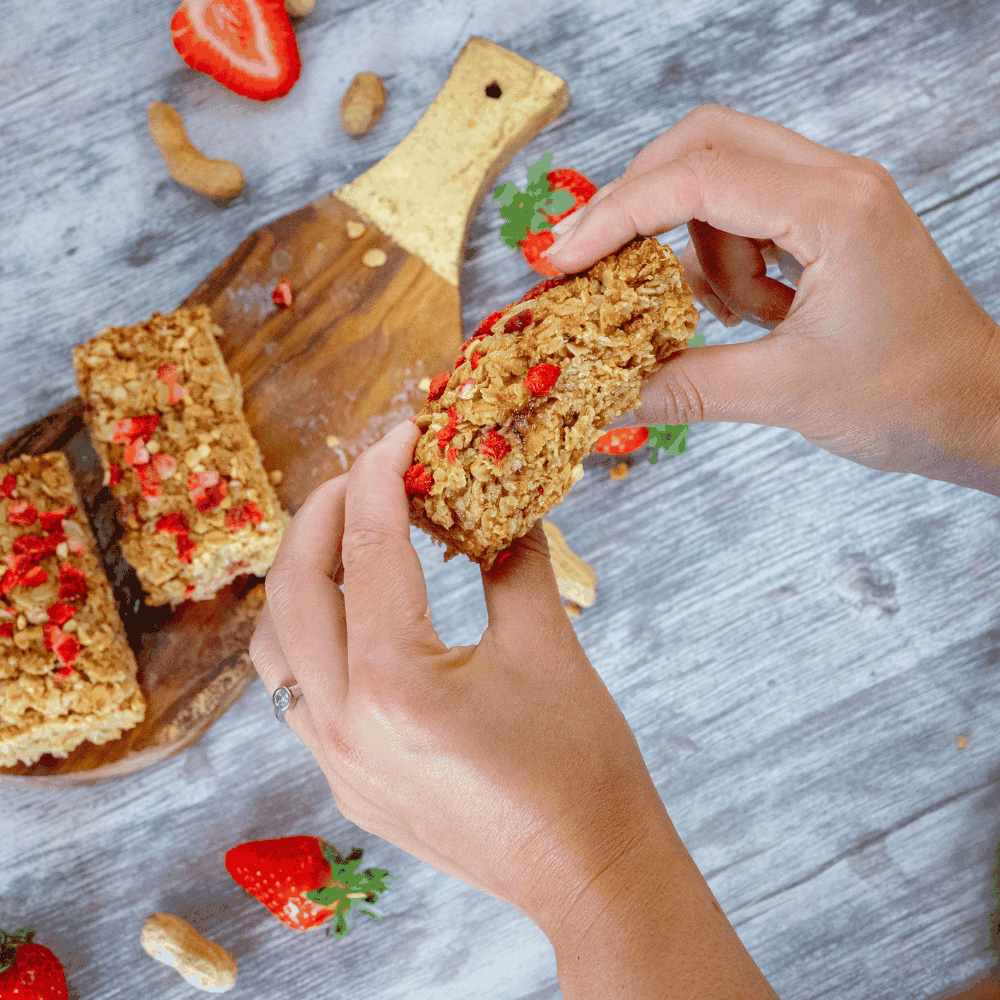Person holding a flapjack bar with strawberries and nuts on a wooden board.