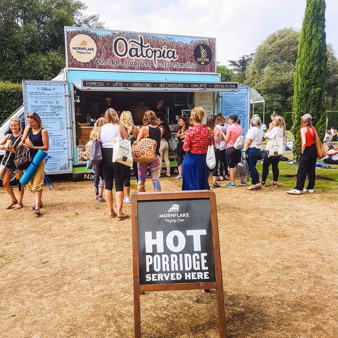 People waiting in line at a food truck named 'Oatopia' with a sign indicating hot porridge is served.