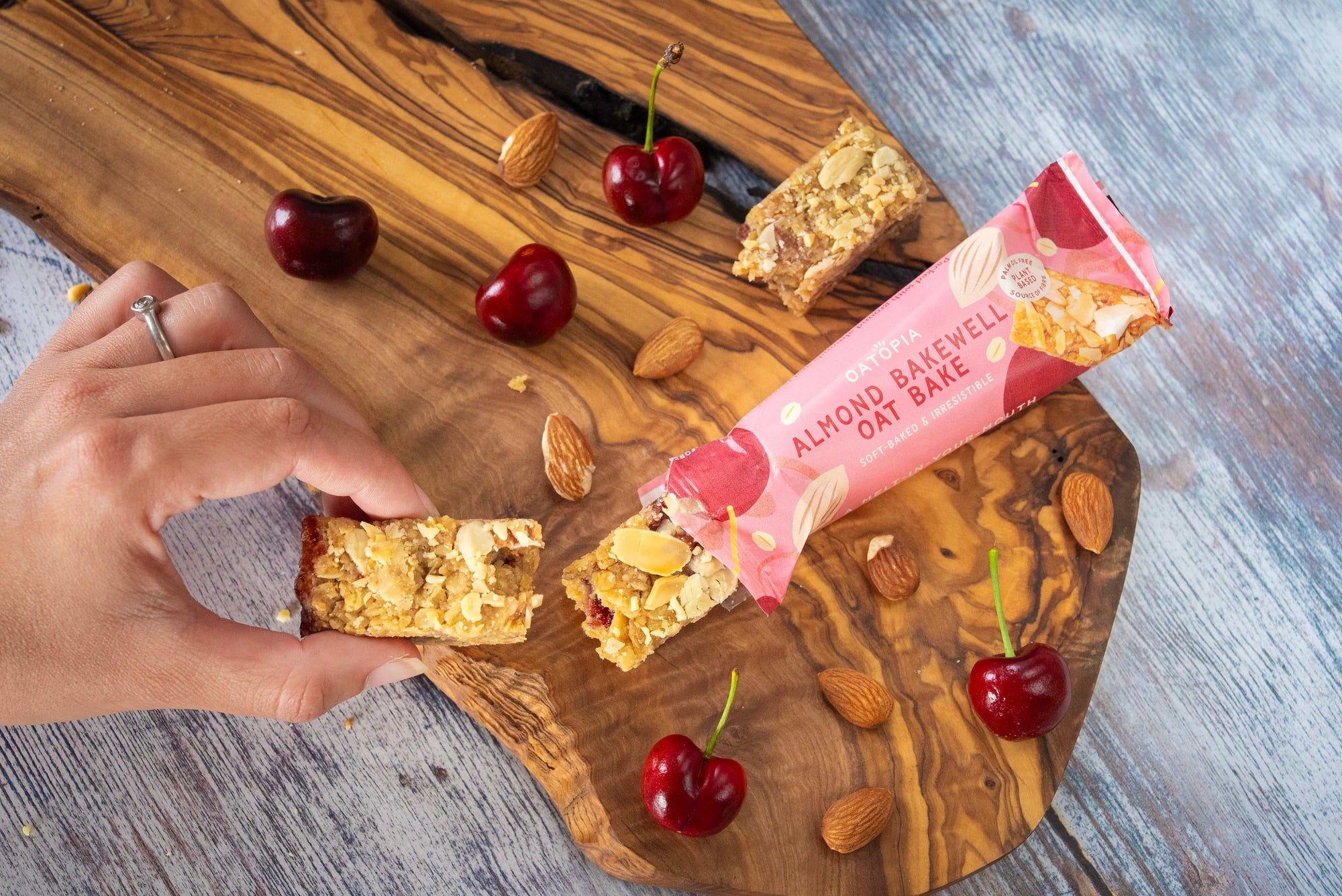 a hand holding half of the oatopia almond bakewell oat bake while the other half is showing the packaging in pink while on the top of the wooden table surround by nuts and fruits. 