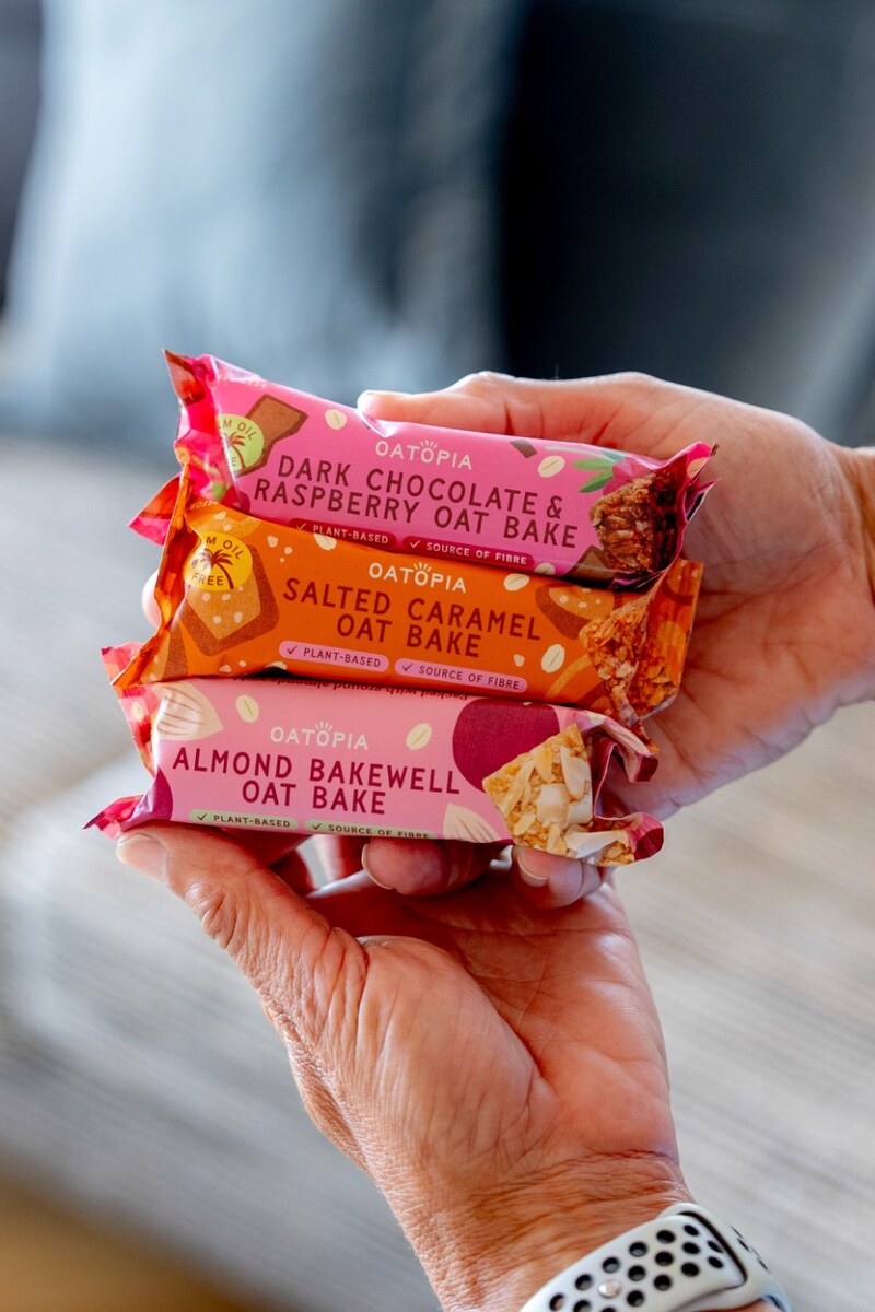 Three Oatopia oat bars in colorful wrappers are held between two hands. The wrappers are pink, orange, and red, with text reading 'OATOPIA' and the names of the three flavors: Dark Chocolate & Raspberry, Salted Caramel, and Almond Bakewell.