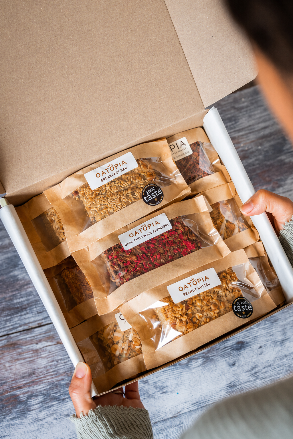 An open cardboard of a box containing four individually wrapped flapjacks on brown paper sleeves, held open by hands over a rustic gray wooden surface. The labels read OATOPIA and include flavors like Oatmeal Chocolate Chip and Oatmeal Peanut Butter.