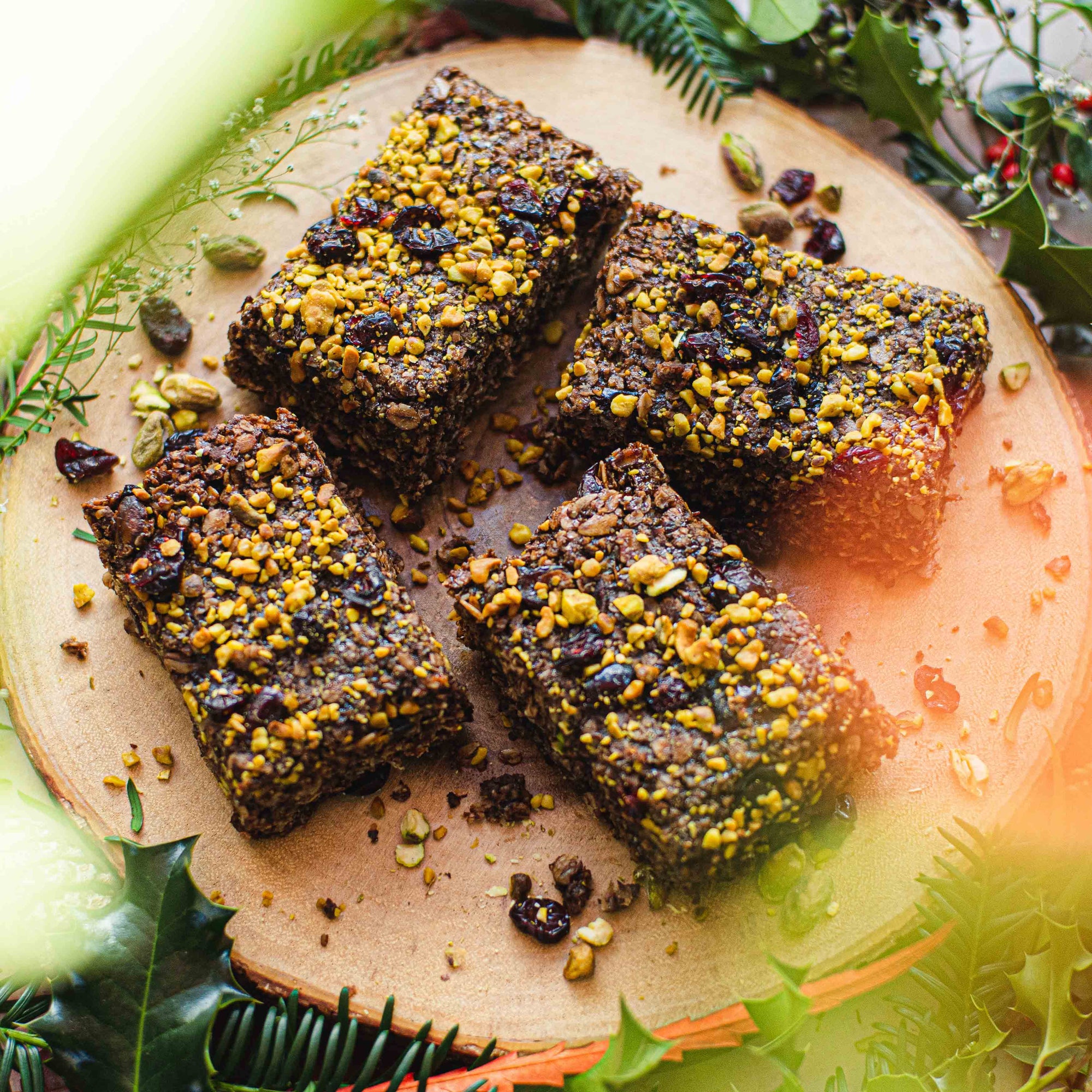 Four square flapjacks with chocolate, pistachio, and cranberry toppings, arranged on a wooden plate with decorative leaves around.